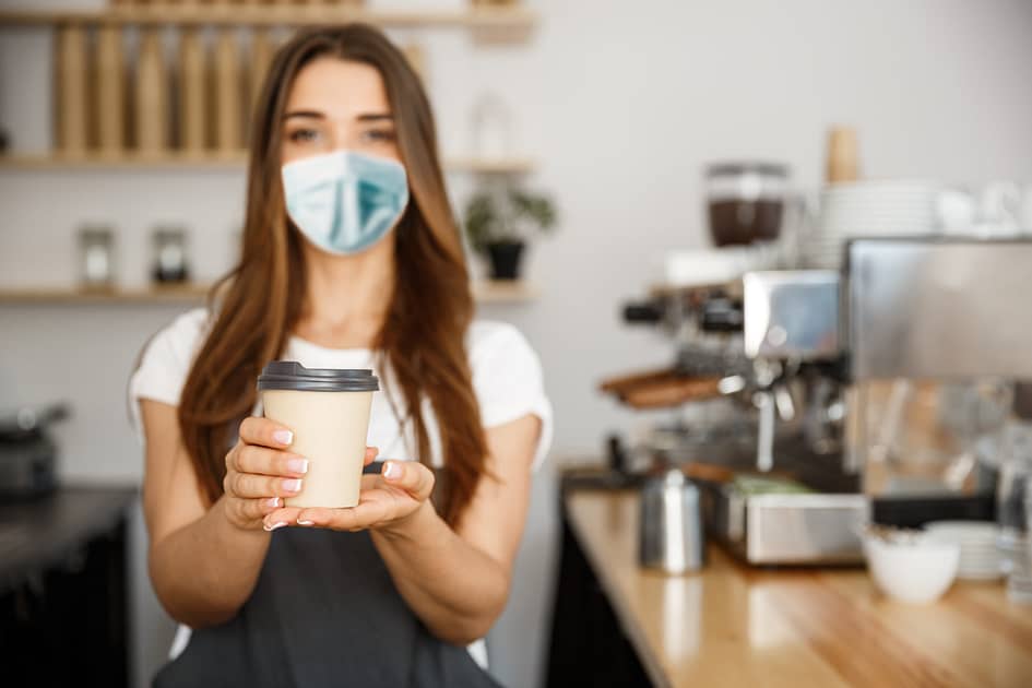 Business Owner Concept - Beautiful Caucasian Barista in face mask offers disposable take away hot coffee at the modern coffee shop.